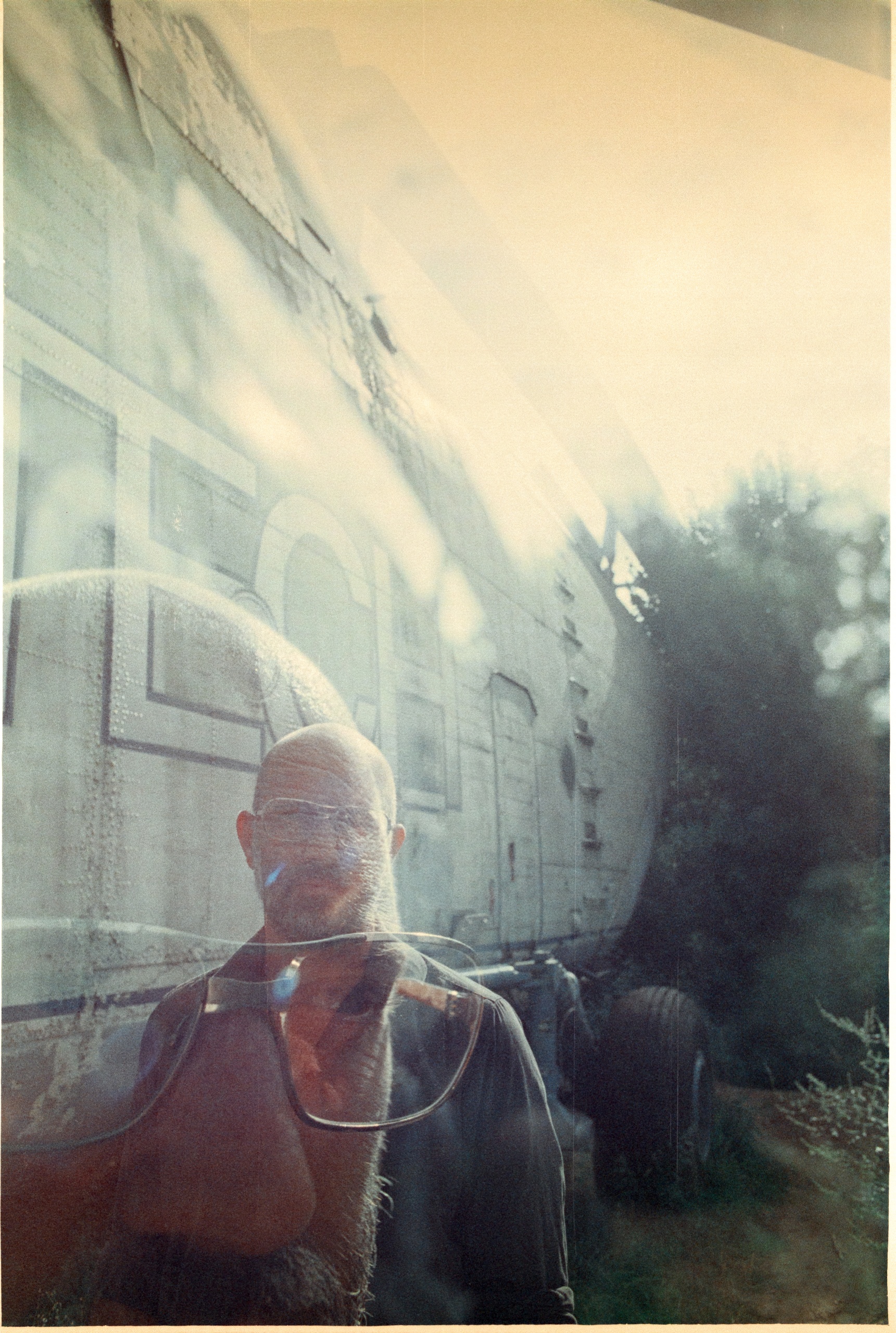 Een man met een baard en bril staat voor een oud vliegtuig in de natuur.