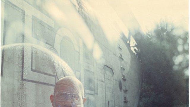 Een man met een baard en bril staat voor een oud vliegtuig in de natuur.