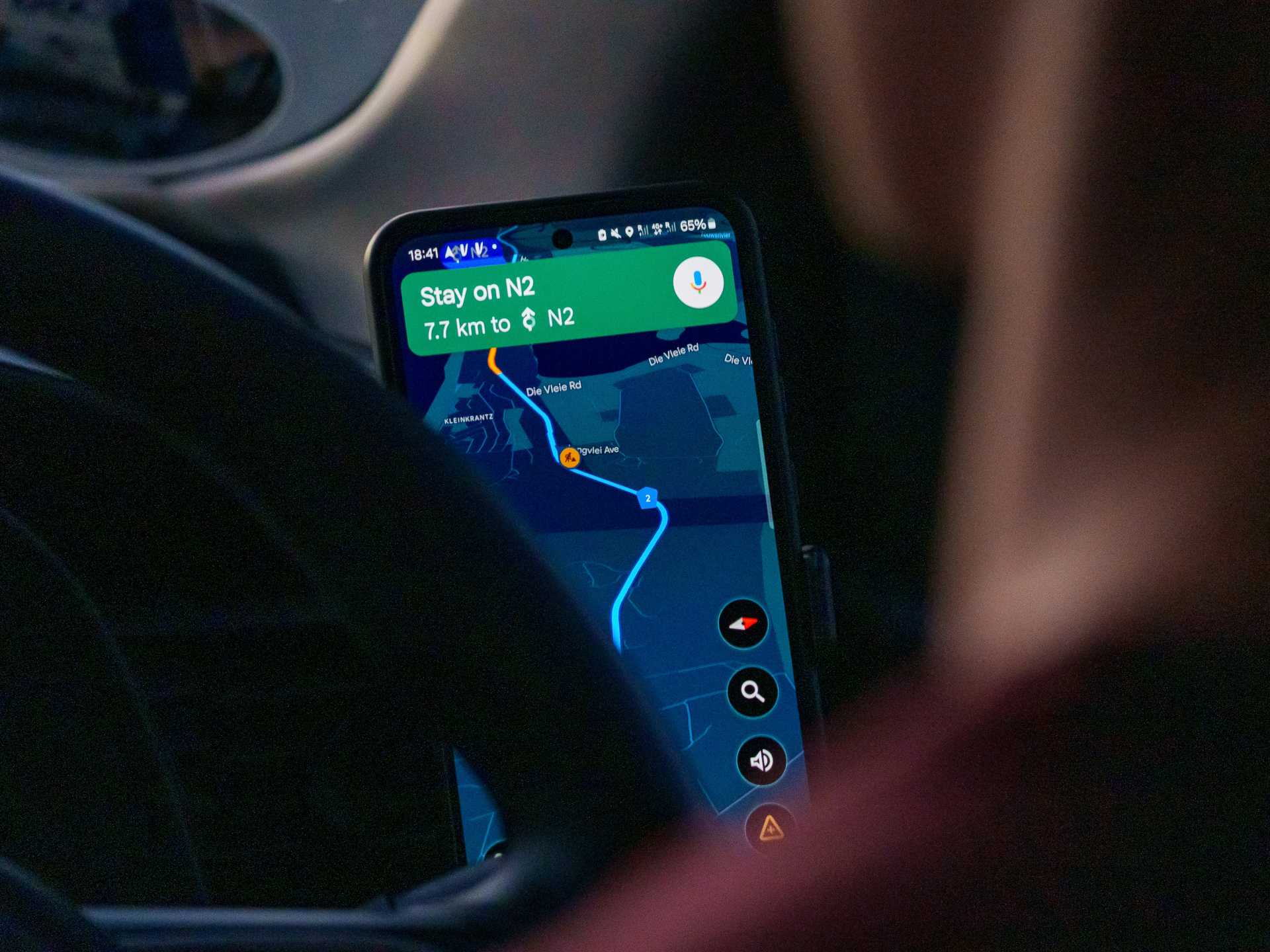 Stuk van een autostuur en persoon zichtbaar, voor een smartphone waardoor Google Maps in nachtmodus een route toont