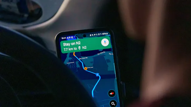 Stuk van een autostuur en persoon zichtbaar, voor een smartphone waardoor Google Maps in nachtmodus een route toont