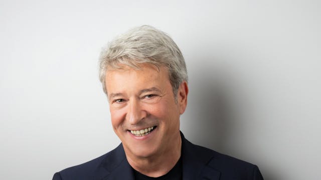 Een glimlachende Garry Hagger met grijs haar, gekleed in een donkerblauw jasje en een zwarte shirt, staat voor een lichte achtergrond. Hij heeft een vriendelijke uitstraling en houdt zijn rechterhand dicht bij zijn borst, terwijl hij naar de camera kijkt.