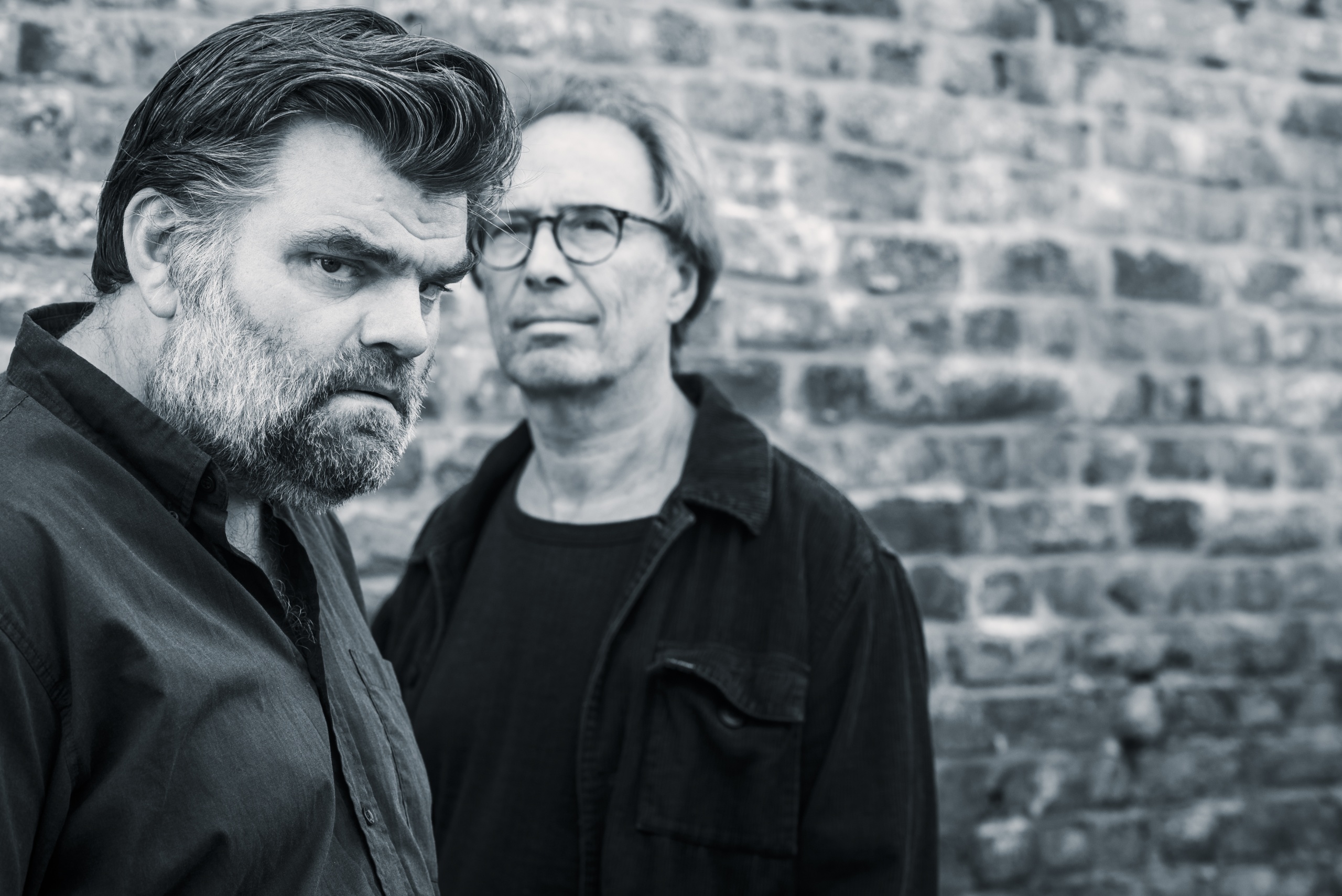 Twee mannen met een intense blik, staand voor een stenen muur. Zwart-wit afbeelding.