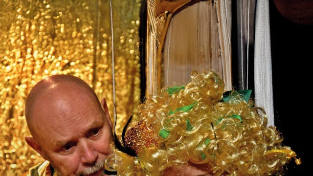 Een man en een vrouw staan dicht bij elkaar. De man kijkt met een geïnteresseerde blik naar de vrouw, die een boos of geïrriteerd gezicht heeft. De vrouw draagt een grote, krullende, gele pruik met groene linten en een witte jas met groene details. De achtergrond is glanzend goudkleurig en geeft een feestelijke sfeer.