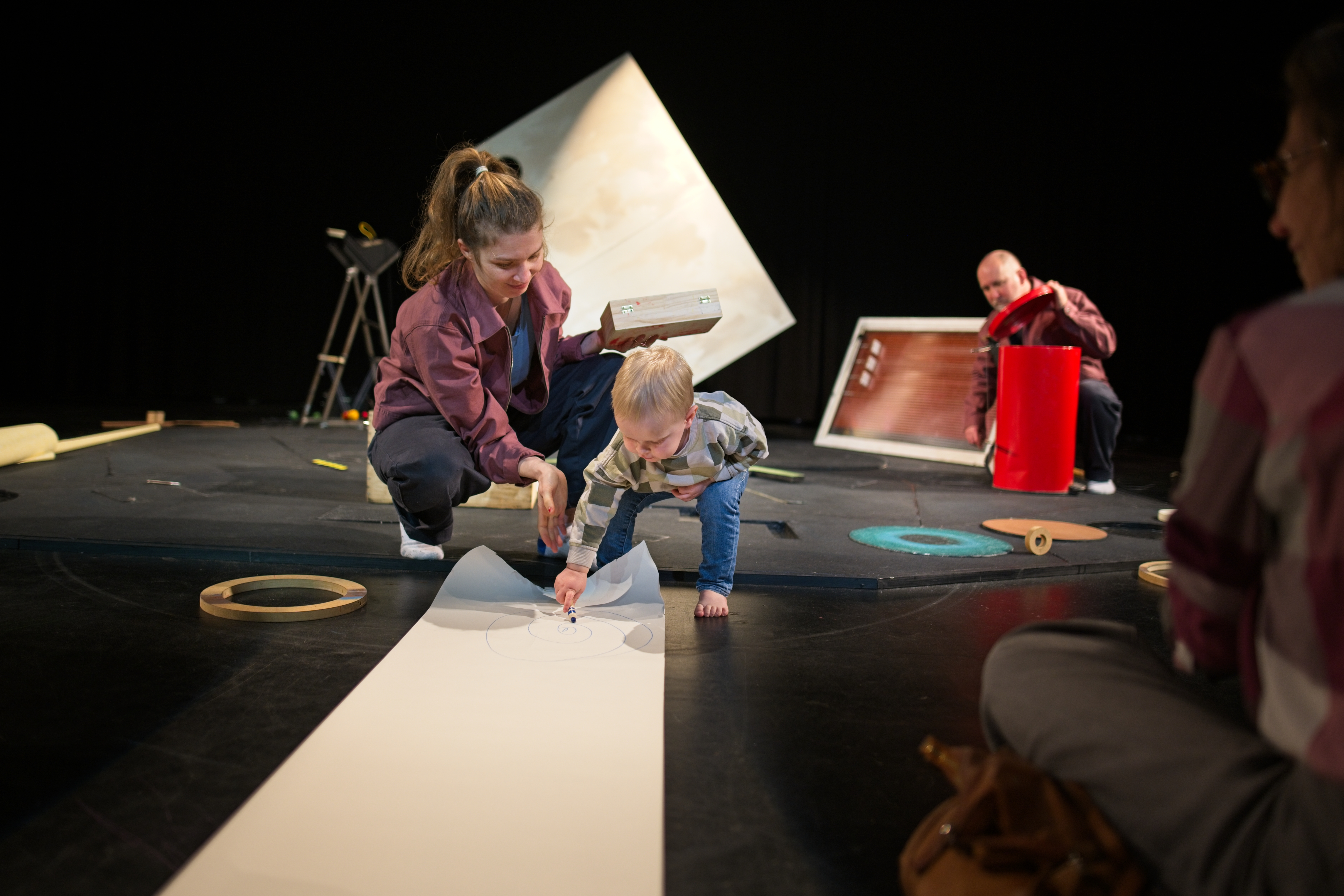 Afbeelding van een persoon die gehurkt een doos vast heeft terwijl een klein kindje op een lang stuk papier tekent.