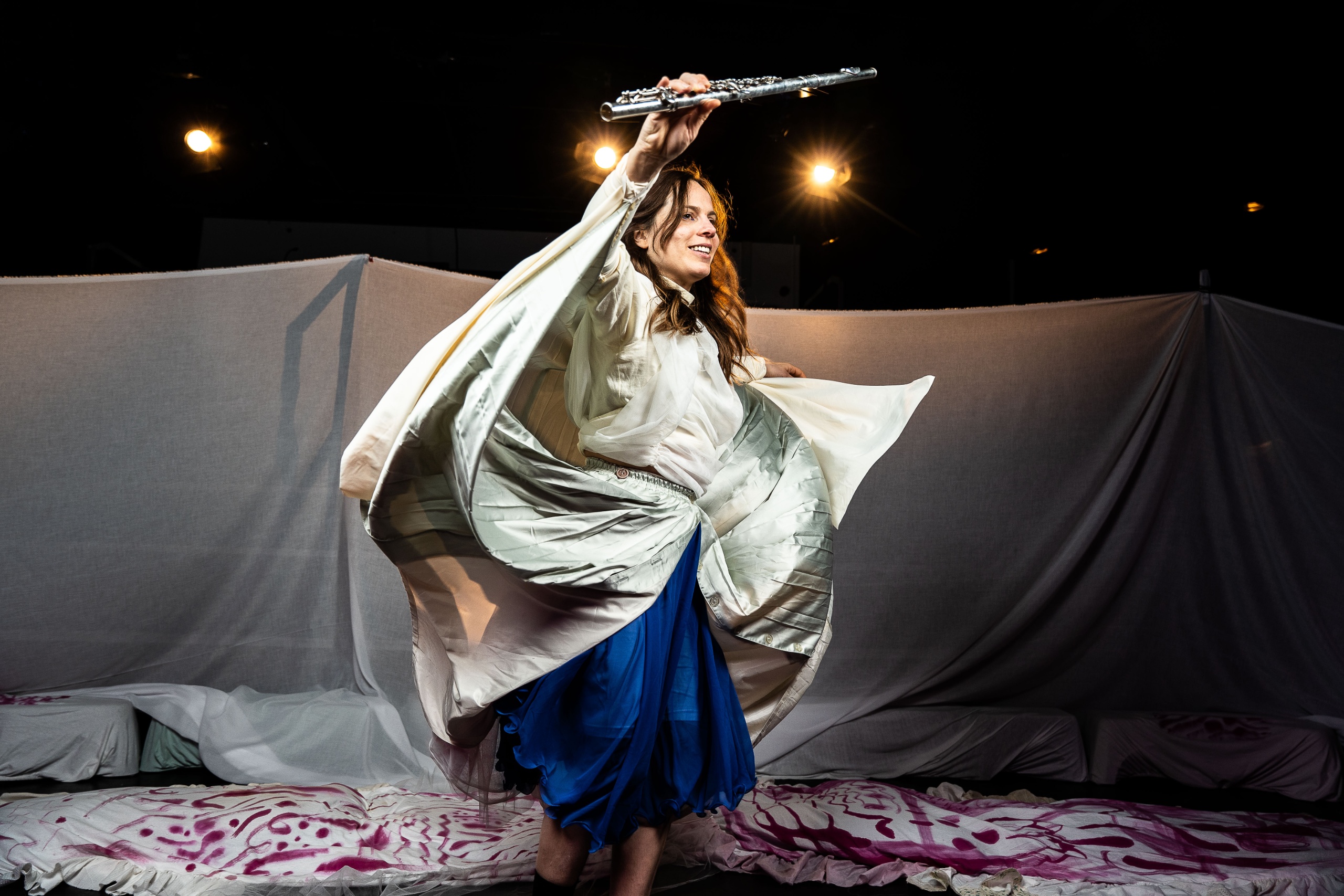 Vrouw in witte en blauwe kleding speelt fluit terwijl ze danst op een podium.