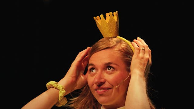 Een vrouw met lichtbruin haar die een gele kroon op haar hoofd plaatst, glimlacht terwijl ze haar handen op haar hoofd legt. Ze draagt een gele outfit met een knuffelbeer op haar borst en een gele haarband. De achtergrond is zwart, waardoor ze duidelijk in de spotlight staat.