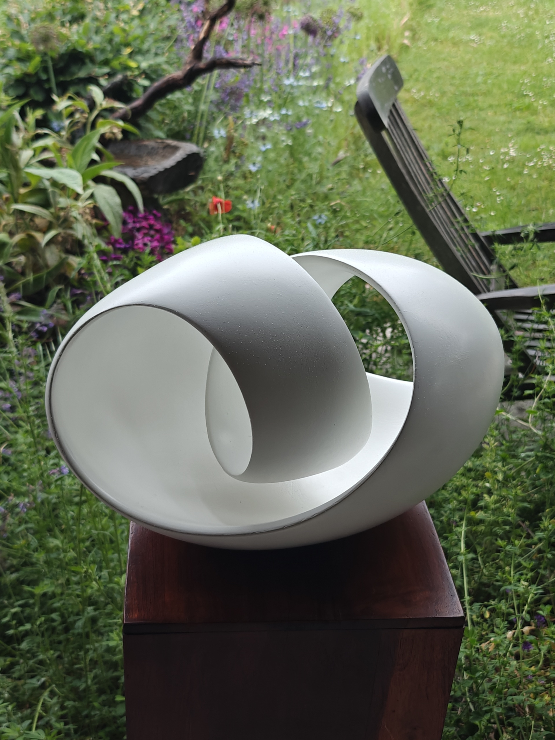 Witte abstracte sculptuur met ronde vormen op houten sokkel in een groene tuin met bloemen en stoel.