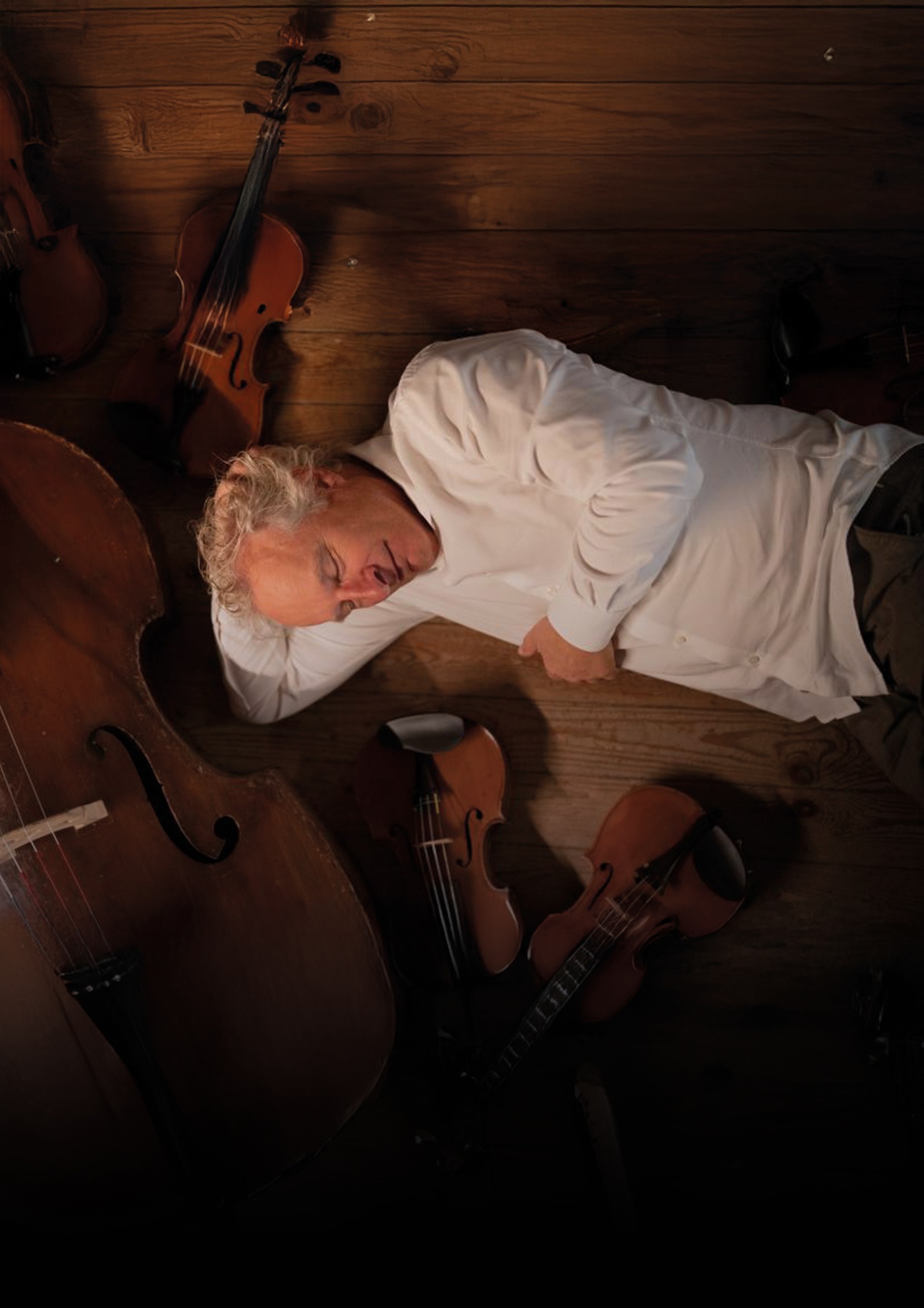 Een man ligt op een houten vloer, slapend of rustend, omringd door violen. Hij draagt een witte blouse en heeft grijs, krullend haar.