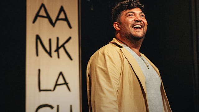 Een man staat op een podium, lachend en kijkend naar boven. Hij draagt een beige jas en een grijs shirt. Achter hem hangt een verticale banner met de tekst "AANKLACHT" geschreven in grote, zwarte letters.