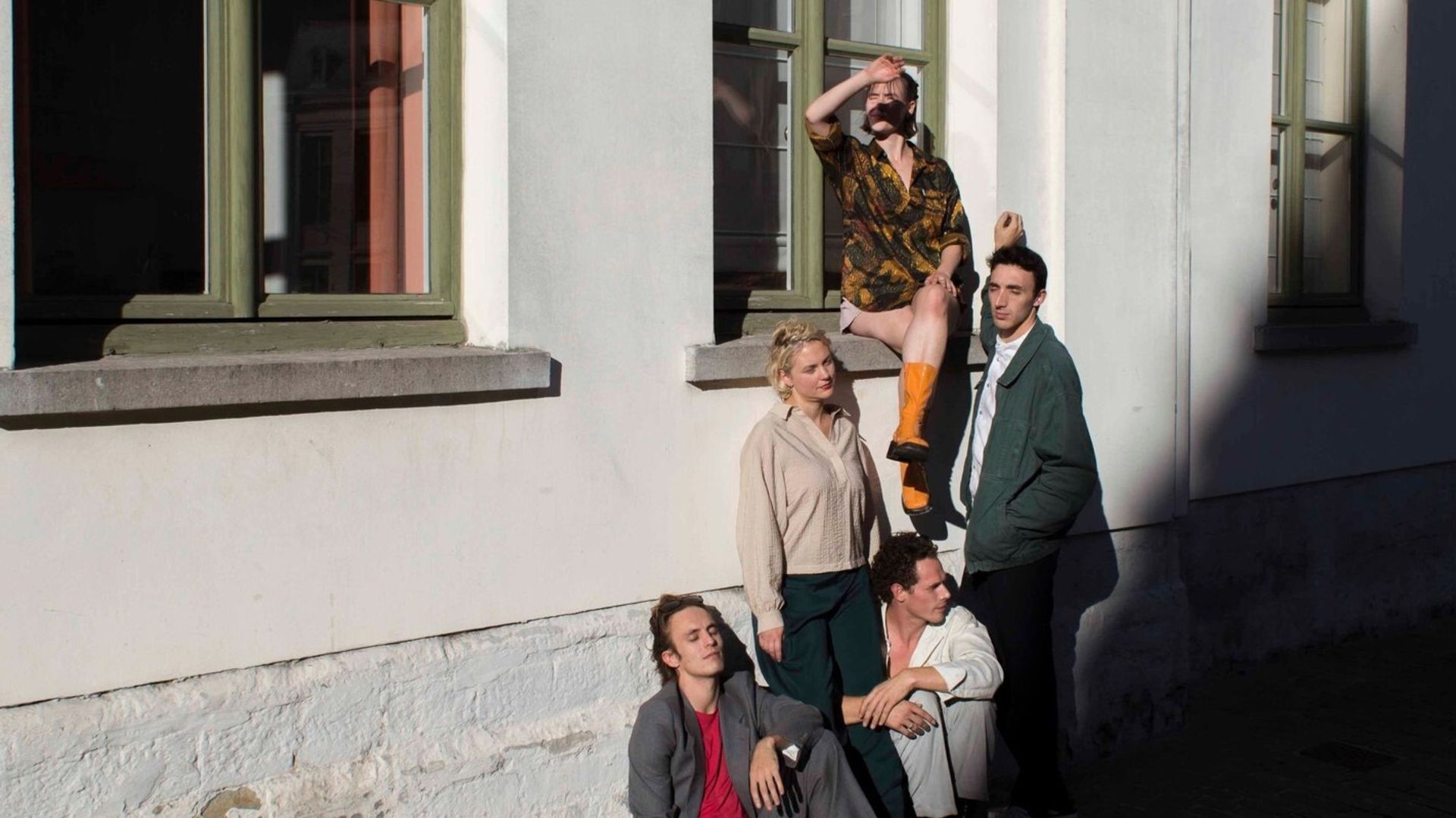 Acteurs zitten en staan tegen beige gevel; één zit op de vensterbank.