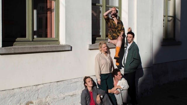 Acteurs zitten en staan tegen beige gevel; één zit op de vensterbank.