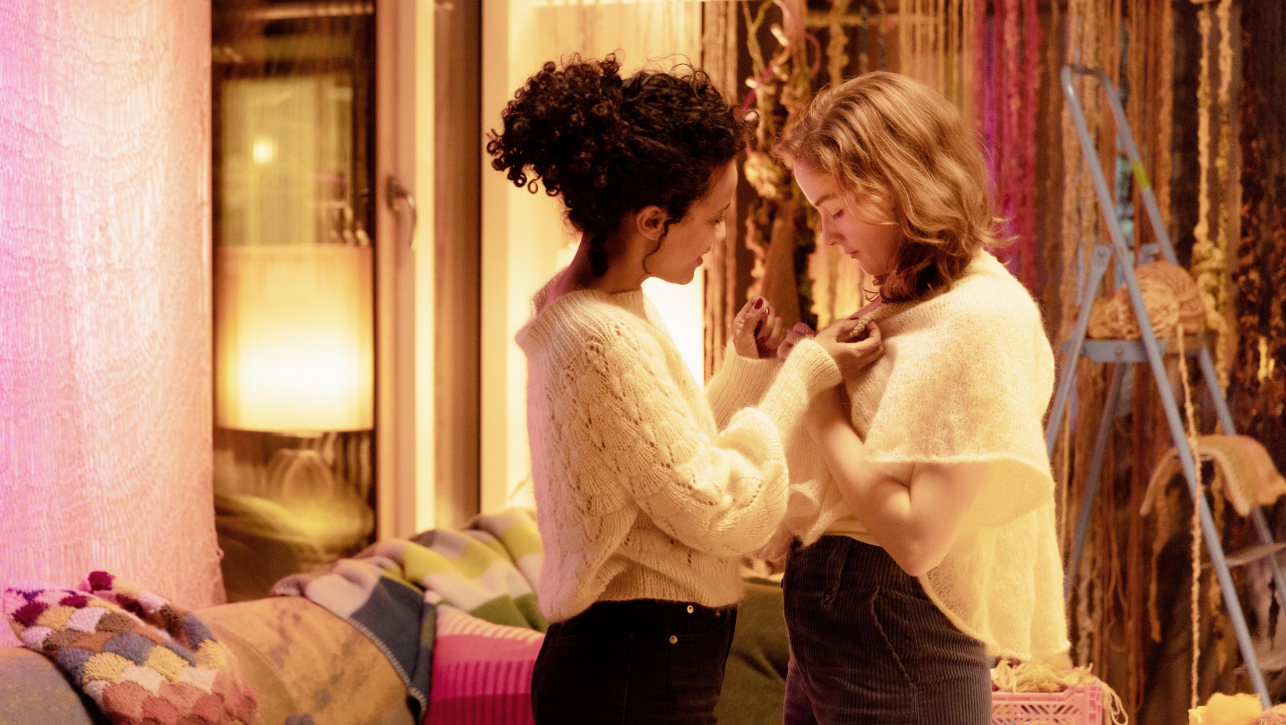 Twee vrouwen helpen elkaar met kleding in een warme, sfeervolle kamer met zachte verlichting.