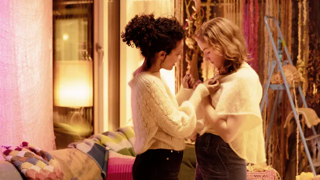 Twee vrouwen helpen elkaar met kleding in een warme, sfeervolle kamer met zachte verlichting.