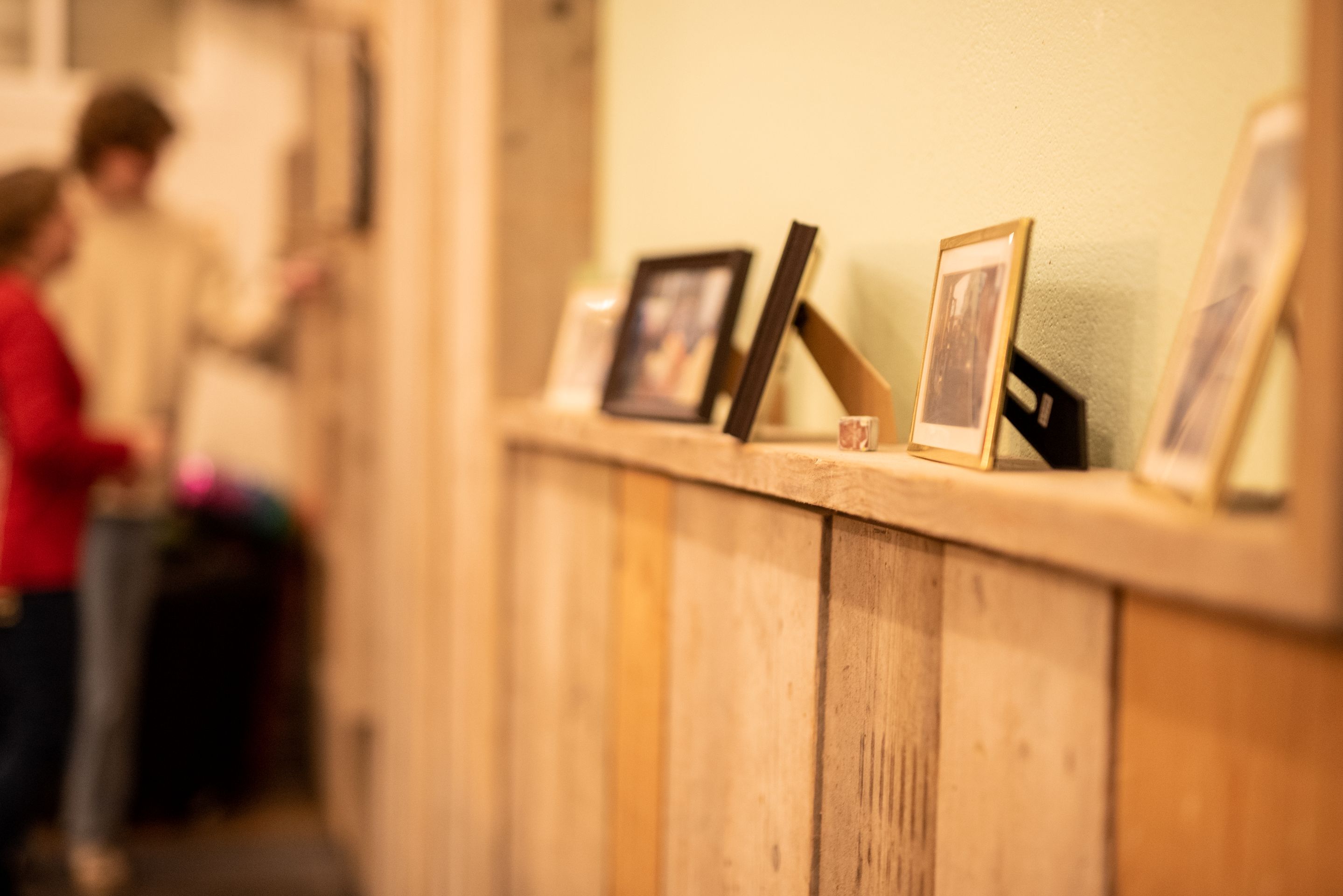 Rij ingelijste foto’s op een houten plank tegen de muur, met twee mensen onscherp op de achtergrond.