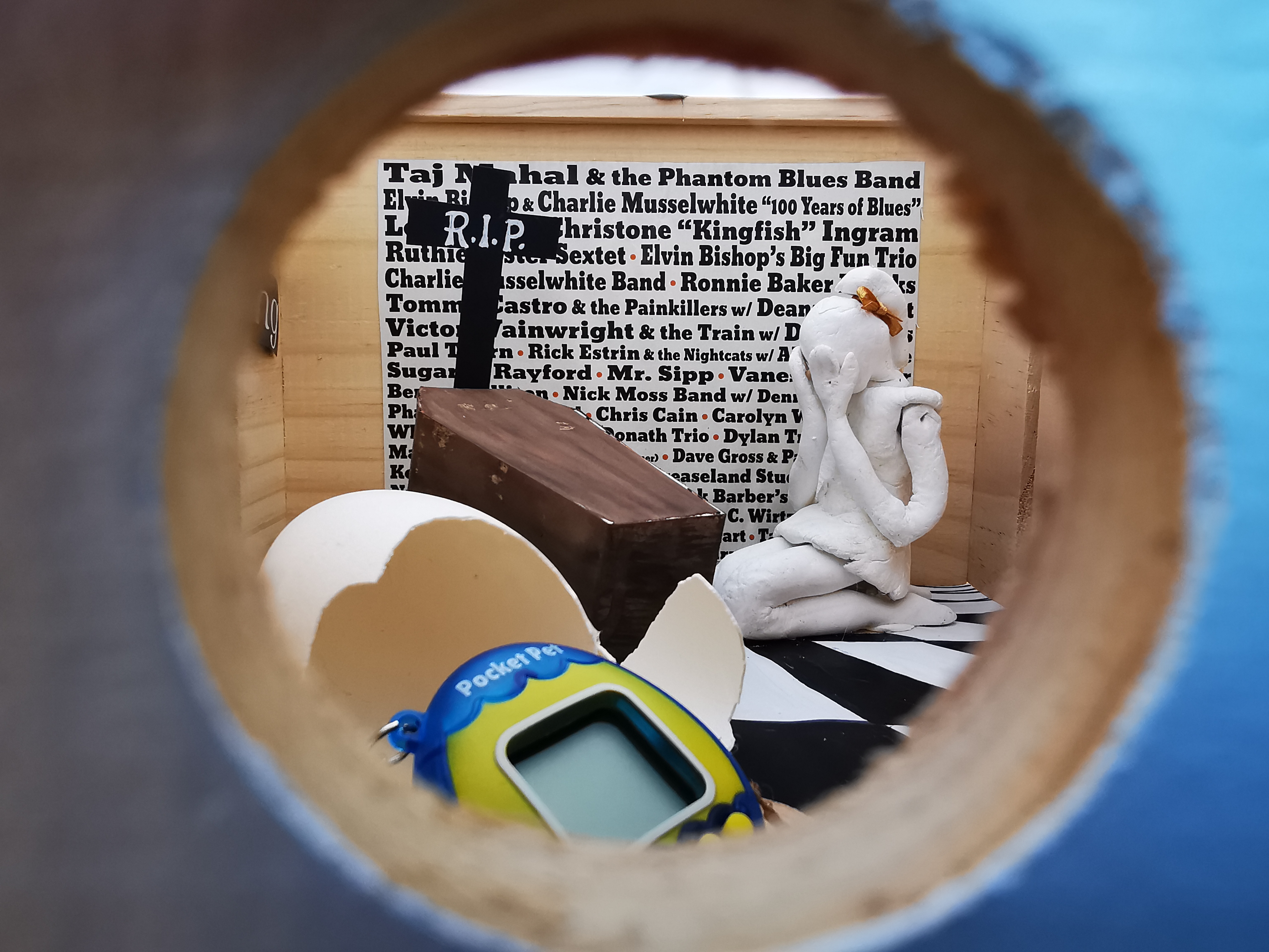 Blik in een kijkdoos met een verbaasd poppetje bij een doodskist, met tekst op de achtergrond