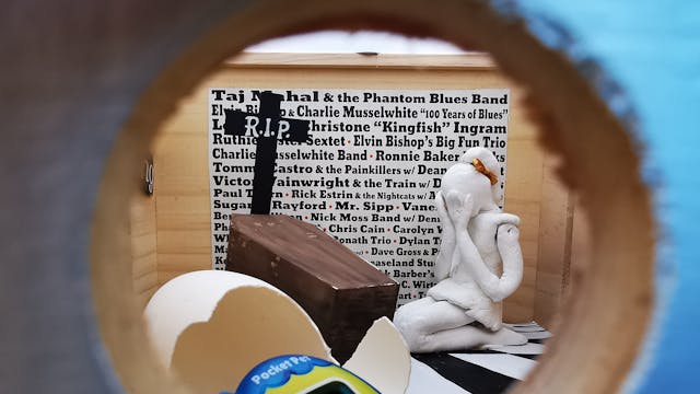 Blik in een kijkdoos met een verbaasd poppetje bij een doodskist, met tekst op de achtergrond