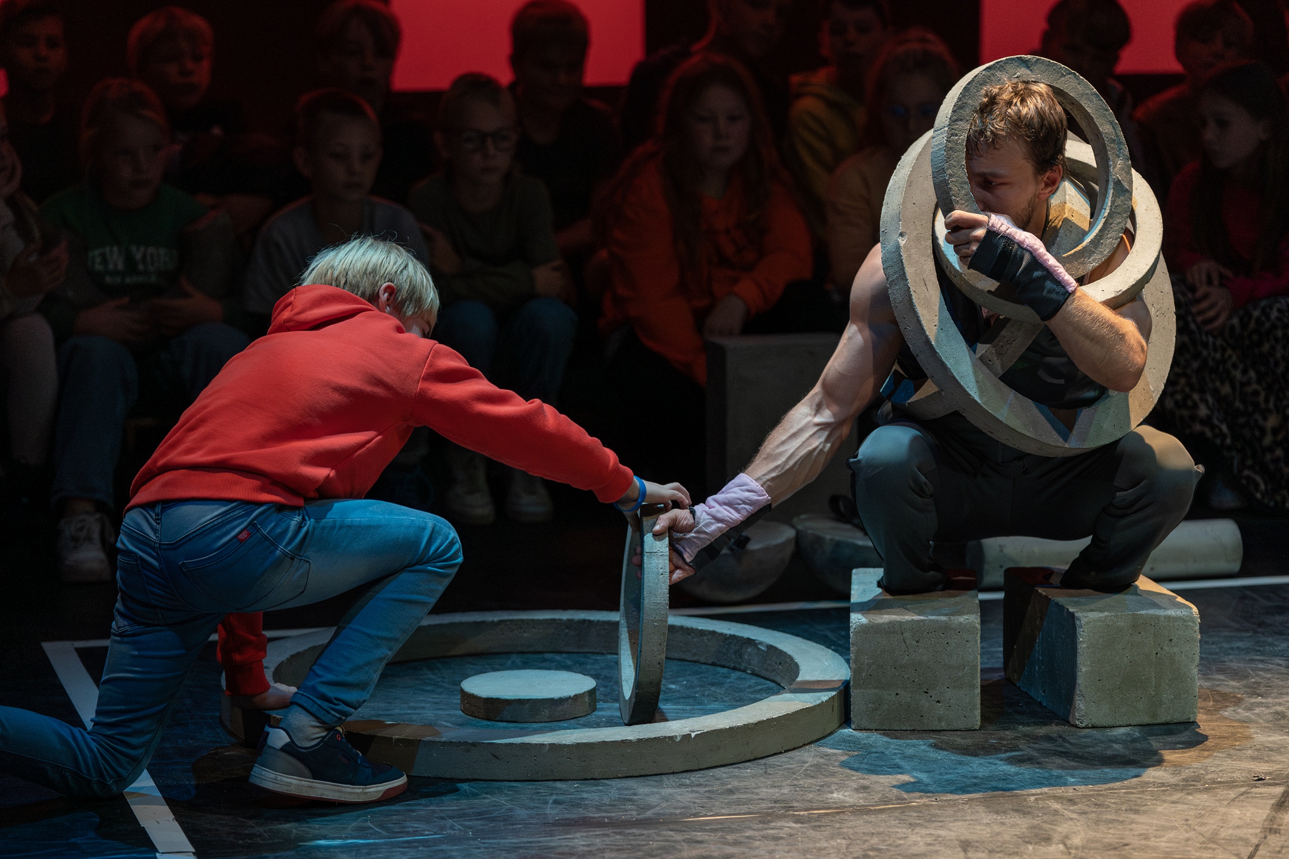 Een jongen in een rode hoodie helpt een krachtatleet met betonnen ringen op het podium.