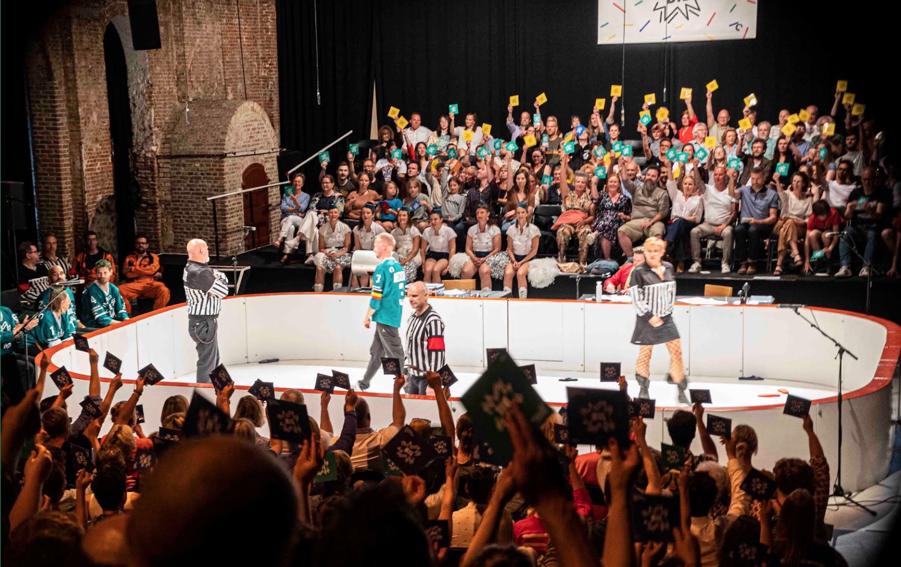 Op deze foto zie je een improvisatietheatervoorstelling die sterk lijkt op een improvisatiematch. De scène is ingericht als een soort sportarena, met een ronde speelvloer en spelers in het midden. Enkele acteurs dragen outfits die doen denken aan scheidsrechters of sporttenues, wat het speelse, competitieve karakter van de voorstelling benadrukt.