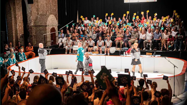 Op deze foto zie je een improvisatietheatervoorstelling die sterk lijkt op een improvisatiematch. De scène is ingericht als een soort sportarena, met een ronde speelvloer en spelers in het midden. Enkele acteurs dragen outfits die doen denken aan scheidsrechters of sporttenues, wat het speelse, competitieve karakter van de voorstelling benadrukt.