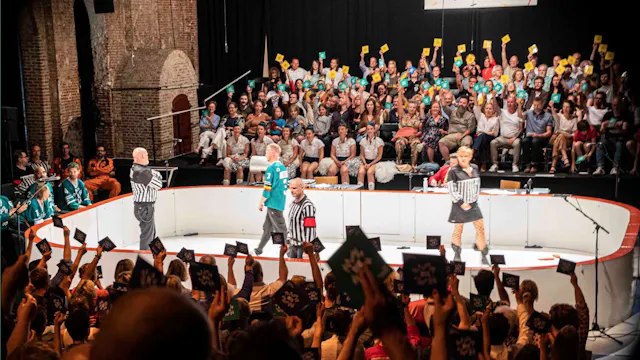 Op deze foto zie je een improvisatietheatervoorstelling die sterk lijkt op een improvisatiematch. De scène is ingericht als een soort sportarena, met een ronde speelvloer en spelers in het midden. Enkele acteurs dragen outfits die doen denken aan scheidsrechters of sporttenues, wat het speelse, competitieve karakter van de voorstelling benadrukt.