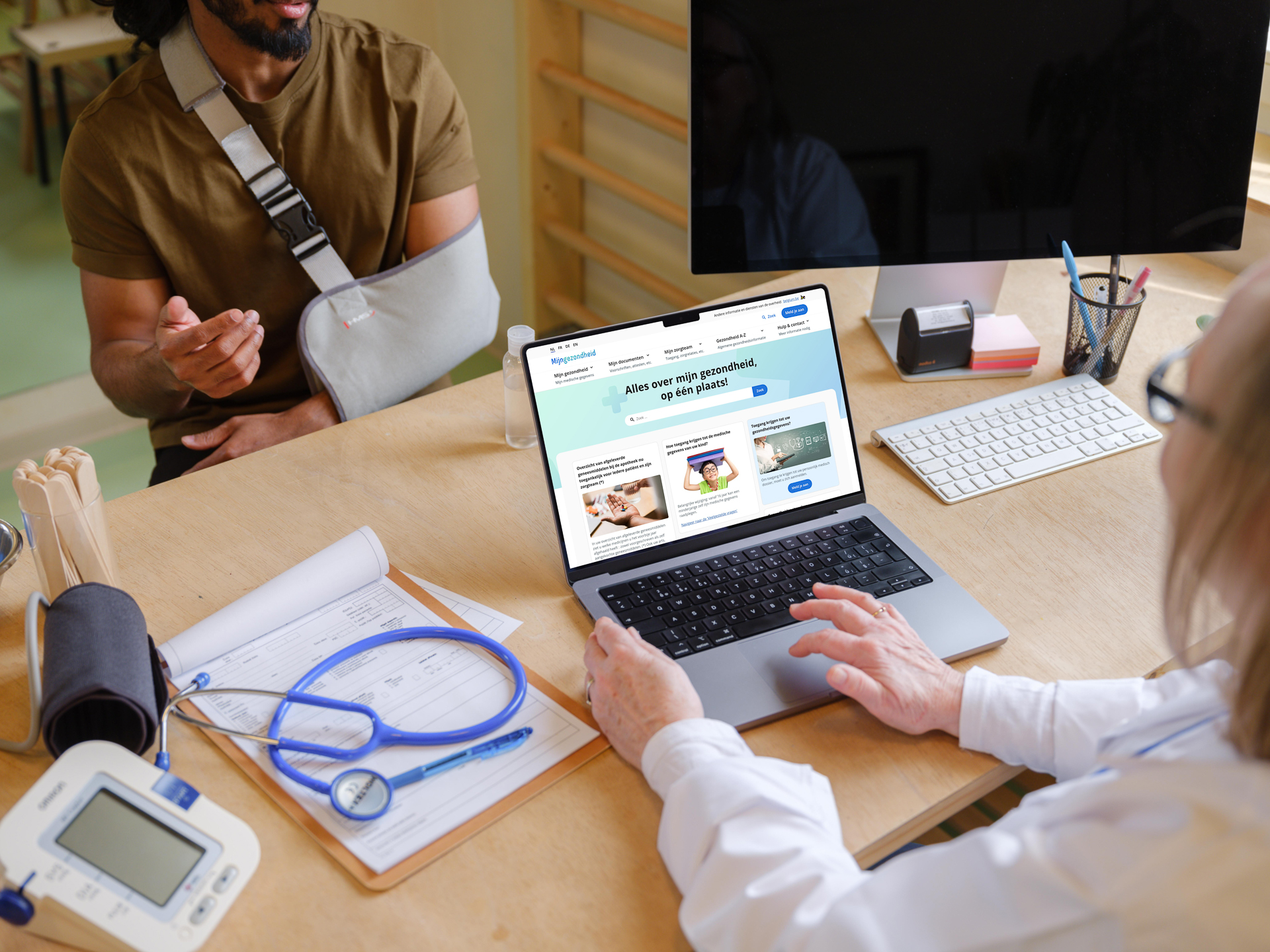 Persoon met arm in verband die bij een zorgverlener zit, die op haar laptop de site mijngzondheid.be heeft open staan