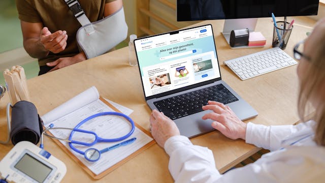 Persoon met arm in verband die bij een zorgverlener zit, die op haar laptop de site mijngzondheid.be heeft open staan