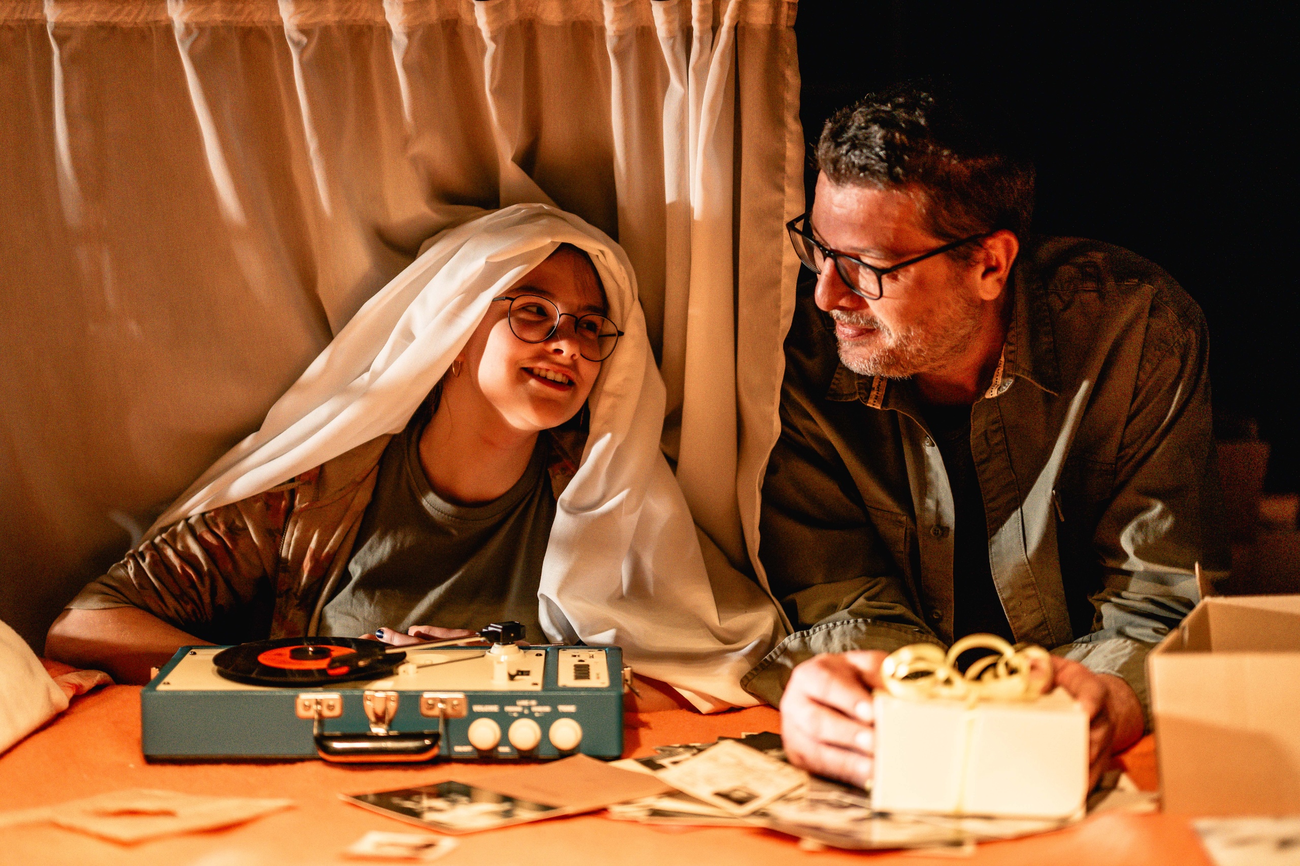 Een jong meisje en een man zitten samen op een bed met een platenspeler en foto's.