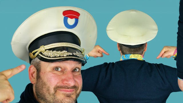 Kapitein Winokio draagt een witte kap met een rood, wit en blauw embleem en een zwarte en gouden militaire pet. Hij wijst naar zijn hoofd met één hand en poseert met een andere man die een grote, witte, ronde hoed draagt. De man met de hoed heeft zijn rug naar de camera en zijn armen gebogen, terwijl hij naar achteren wijst. De onderste helft van de afbeelding toont een persoon in witte broek en gele schoenen, met een hand die een duim naar beneden geeft. De achtergrond is lichtblauw.