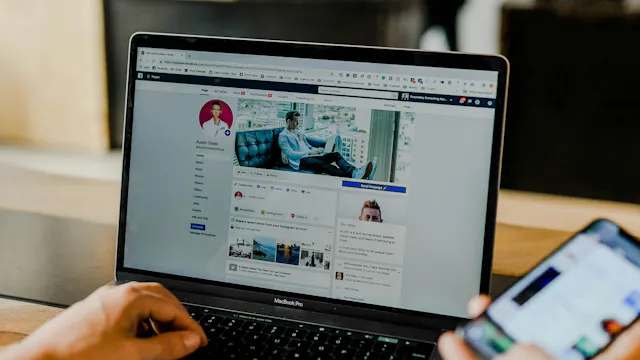 Laptop waar Facebook-pagina open staat, ene hand op toetsenbord, andere hand houdt smartphone vast