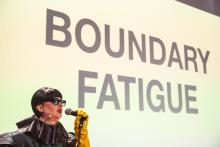 Afbeelding van persoon met microfoon vast voor een groot doek met tekst op geprojecteerd.