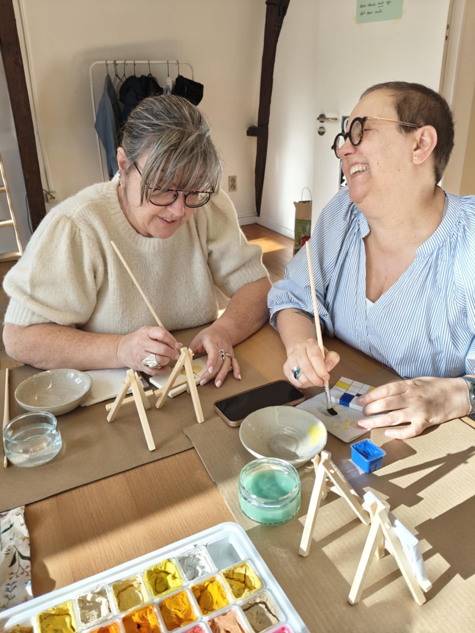 2 vrouwen zijn samen creatief aan het knutselen in een atelier. Je ziet verf en kleine canvassen. Er wordt gelachen