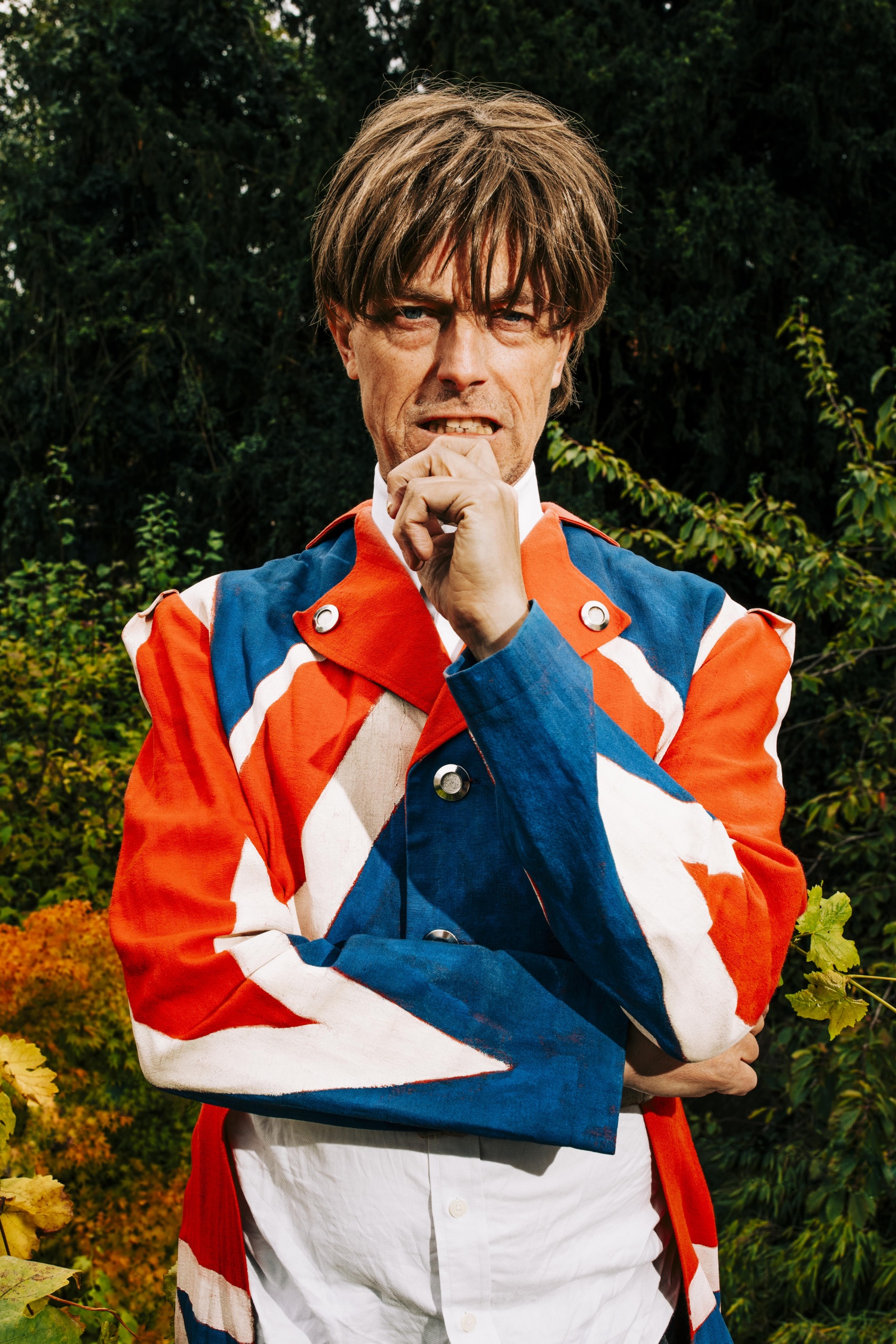 Man in jas met Britse vlag, hand bij mond, staand voor groen struikgewas.