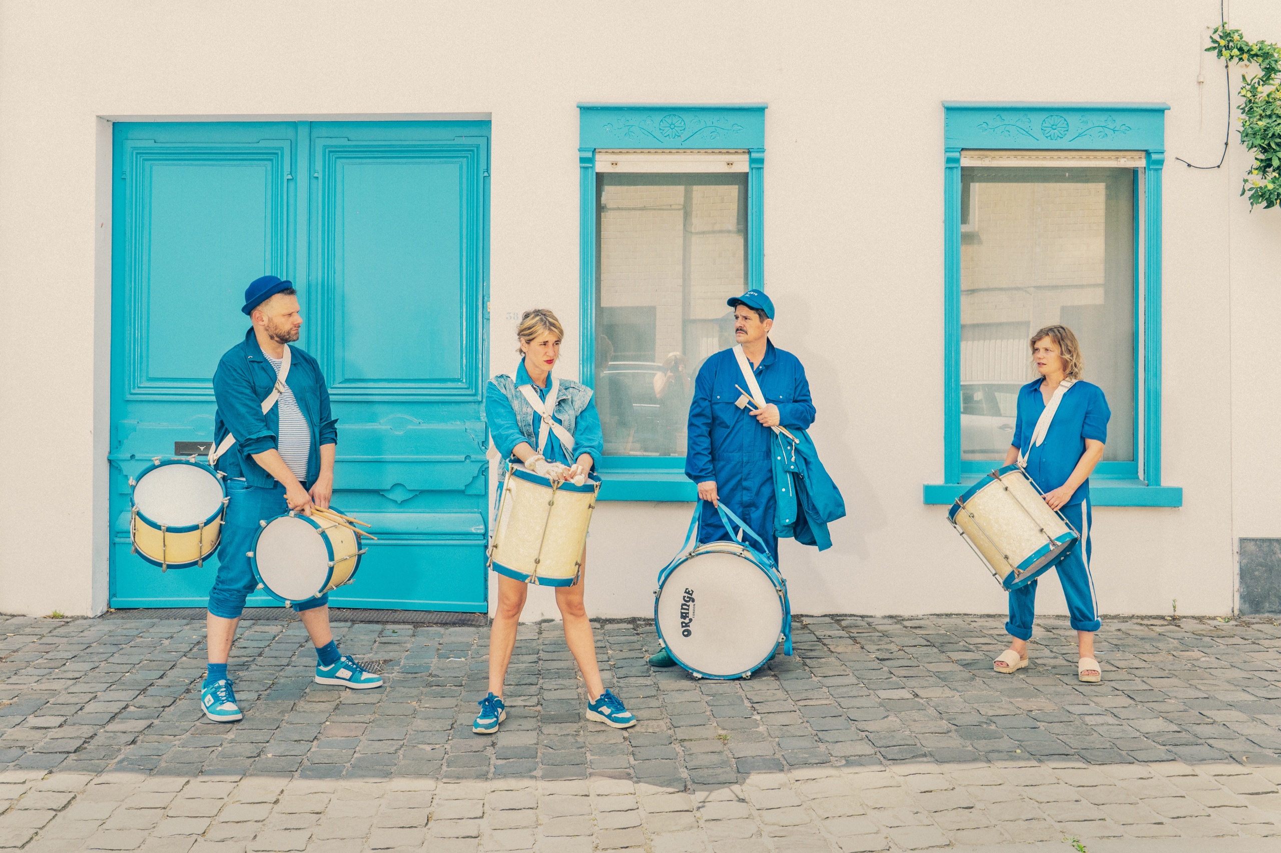 Vier mensen in blauwe kleding staan buiten voor een wit gebouw met felblauwe deur en ramen. Ze dragen elk een trommel en drumstokken, klaar om te spelen. De straat is geplaveid met kasseien.