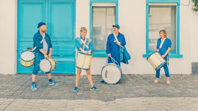 Vier mensen in blauwe kleding staan buiten voor een wit gebouw met felblauwe deur en ramen. Ze dragen elk een trommel en drumstokken, klaar om te spelen. De straat is geplaveid met kasseien.