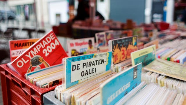 Op de voorgrond bakken vol met strips, waarvan er enkele bovenuit steken. Op de achtergrond staat iemand aan het kraampje tussen boeken of strips te snuisteren.