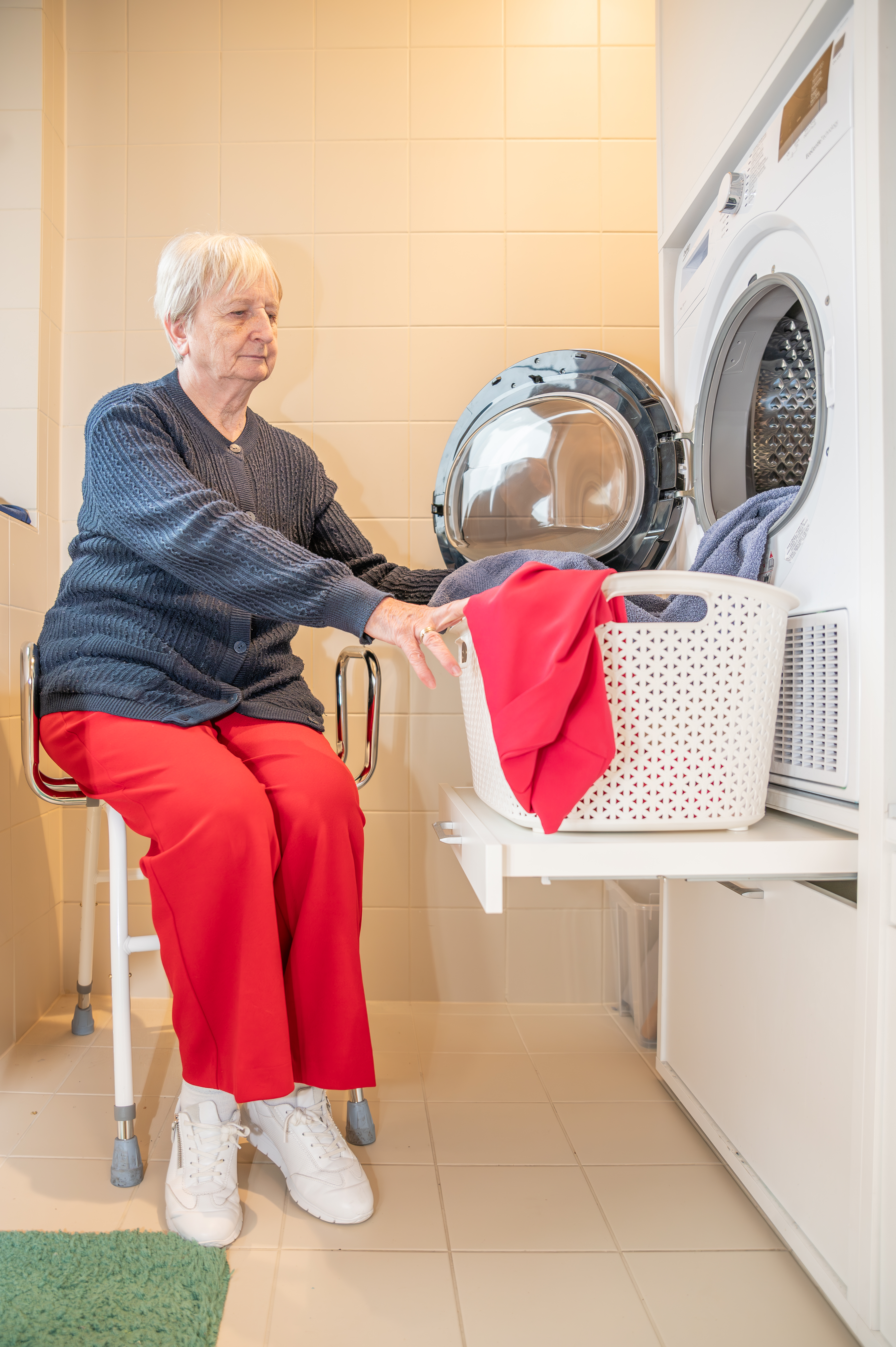 Levenslang wonen. Vrouw zit op aangepaste stoel aan een wasmachine met een speciale legger om de wasmand op te plaatsen.