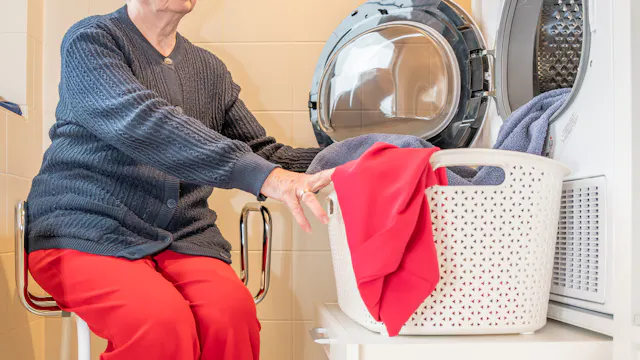 Levenslang wonen. Vrouw zit op aangepaste stoel aan een wasmachine met een speciale legger om de wasmand op te plaatsen.