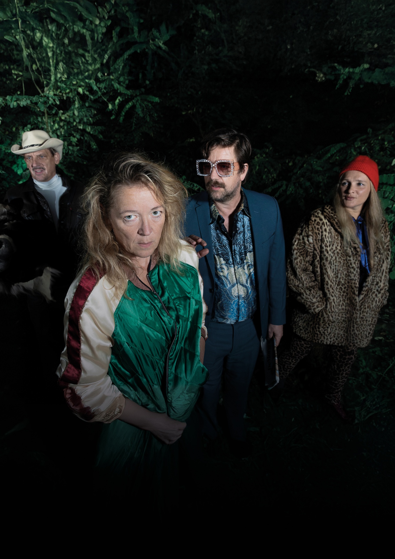 Een groep mensen staat in een bos, gekleed in opvallende en kleurrijke kleding. De vrouw in de voorgrond draagt een groene en beige windjack en kijkt serieus naar de camera. Naast haar staat een man met een blauwe jas, een opvallende bril met grote glazen en een getailleerd overhemd met een blauw patroon. Aan de rechterkant staat een vrouw in een leopardprint jas en een rode muts, die naar de groep kijkt. In de achtergrond is een man te zien met een cowboyhoed en een zwart jack, die naar de zijkant kijkt. De sfeer is enigszins excentriek en de setting lijkt nachtelijk of in een donkere omgeving.