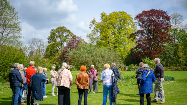 Gids vertelt in het arboretum aan een groep bezoekers die vrolijk gekleurde kleding dragen