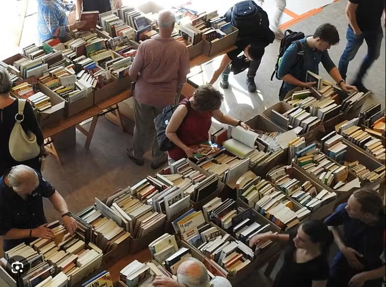 Een overzicht van een boekenmarkt met meerdere tafels vol met boeken. Verschillende mensen bladeren door de boeken en kijken of ze iets interessants kunnen vinden. De setting lijkt een drukke, gezellige plek waar boekenliefhebbers samenkomen.