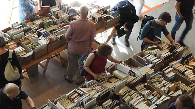 Een overzicht van een boekenmarkt met meerdere tafels vol met boeken. Verschillende mensen bladeren door de boeken en kijken of ze iets interessants kunnen vinden. De setting lijkt een drukke, gezellige plek waar boekenliefhebbers samenkomen.