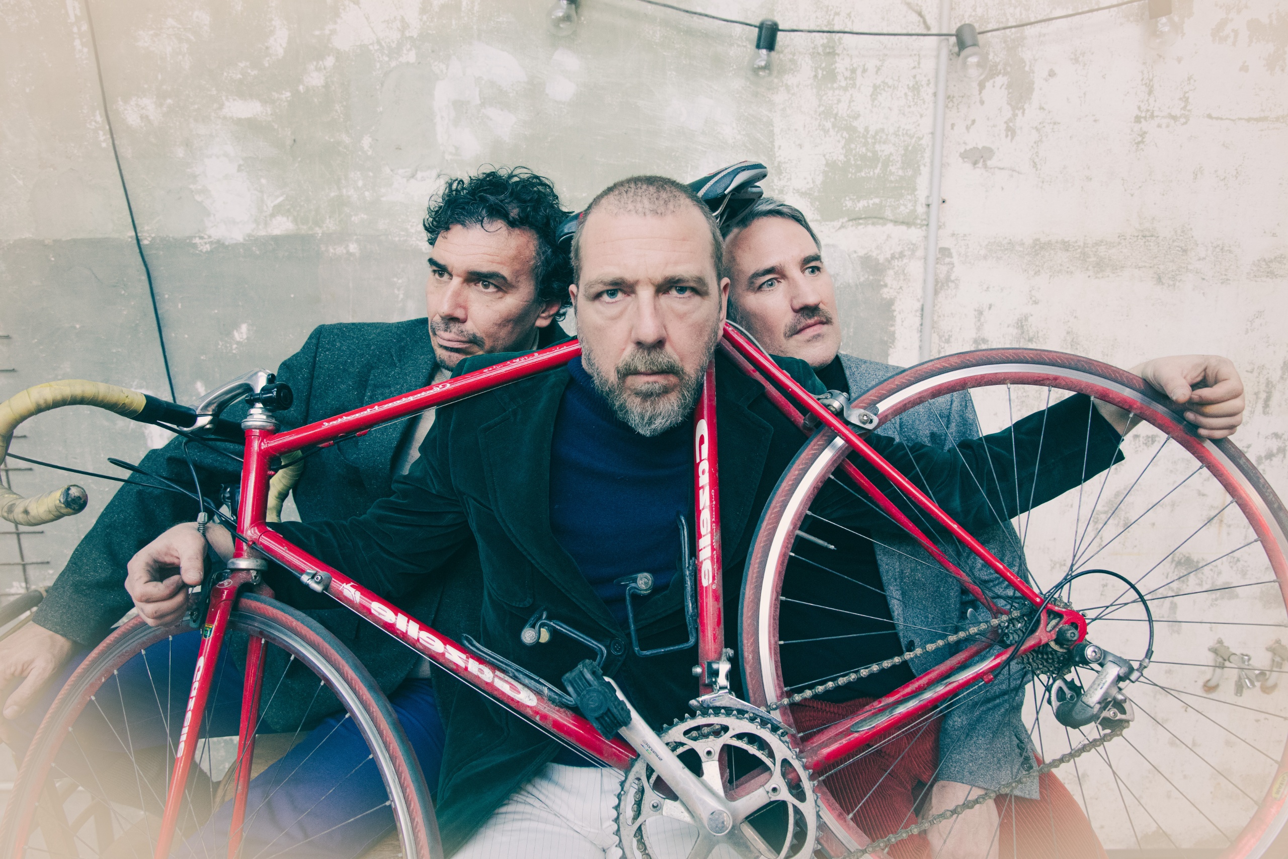 Drie mannen poseren met een rode fiets in een industriële setting.