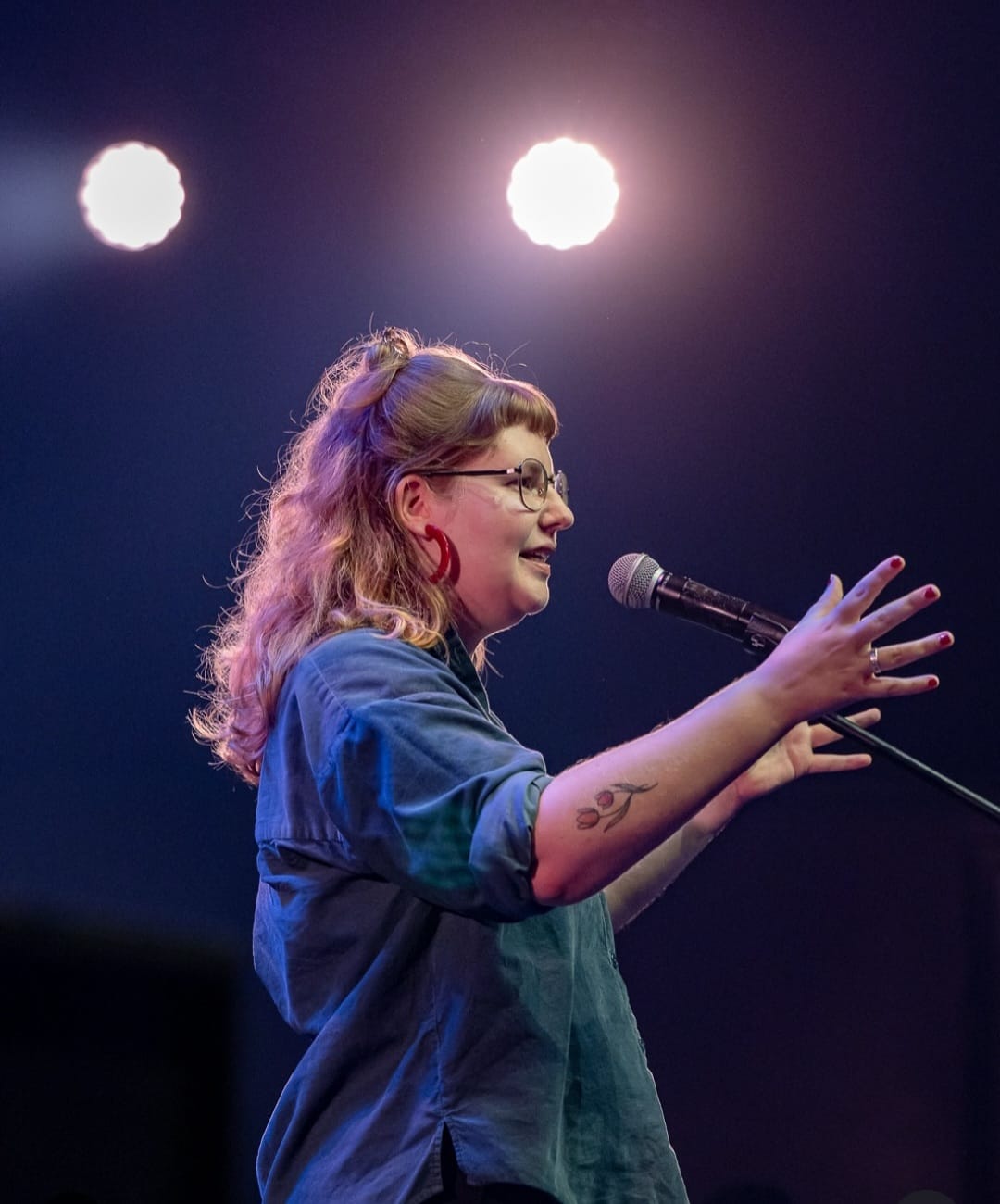 Vrouw met bril en tatoeage spreekt enthousiast in microfoon op podium met fel licht op achtergrond.