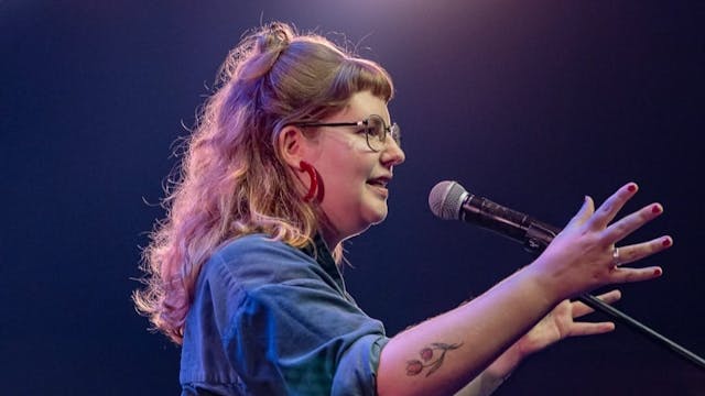 Vrouw met bril en tatoeage spreekt enthousiast in microfoon op podium met fel licht op achtergrond.
