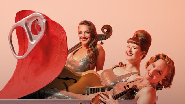 Drie vrouwen in glitterende outfits zitten in een grote, visvormige bak met een visafbeelding aan de zijkant. Ze houden muziekinstrumenten vast en lachen naar de camera. Eén vrouw houdt een gitaar, een andere een viool, en de derde vrouw zit achter een grote, rode blikjesvormige prop. De achtergrond is effen lichtroze.