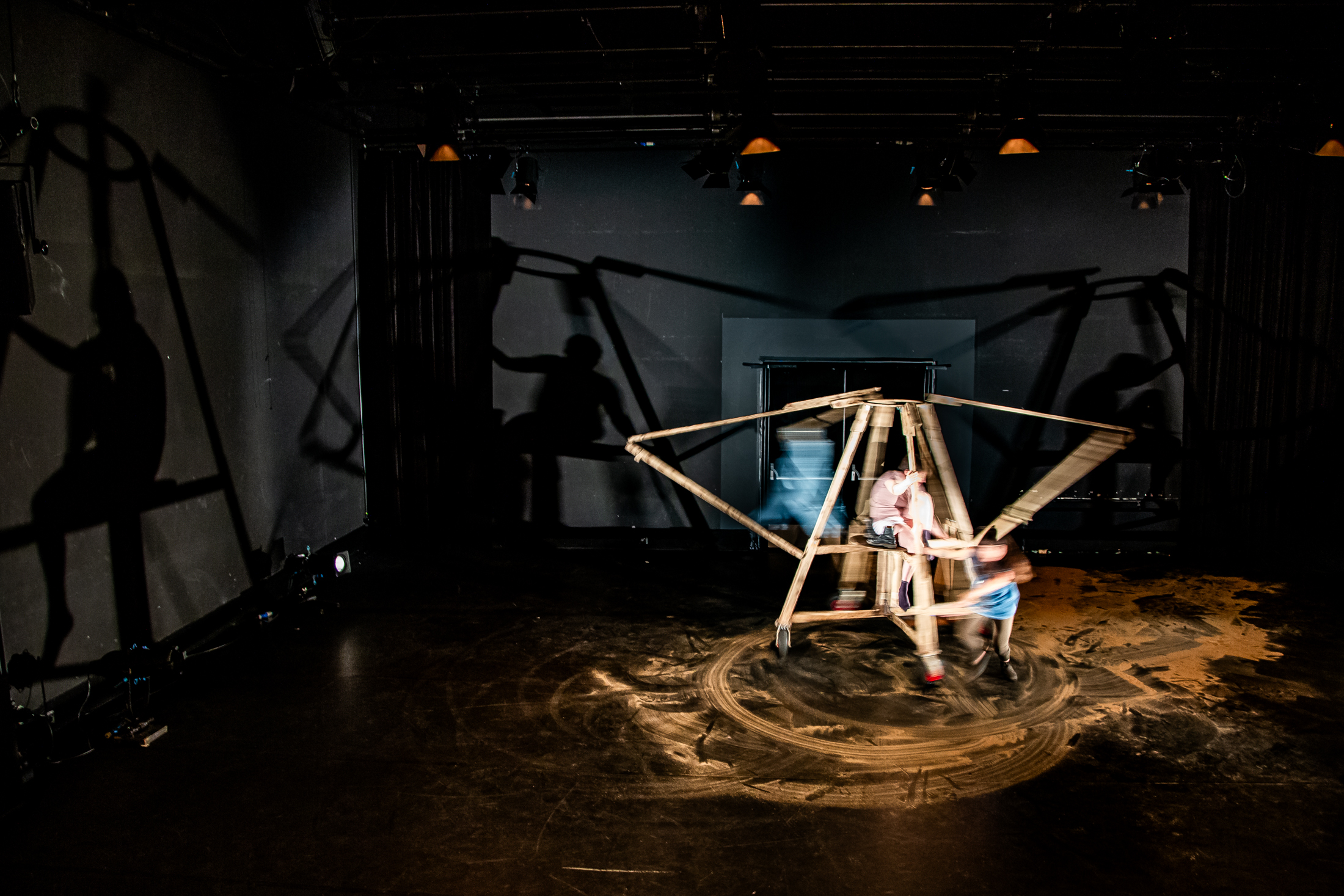 Foto uit de voorstelling Oef!. Op het podium staat een houten installatie, de schaduwen ervan zijn rondom rond op de muren te zien.