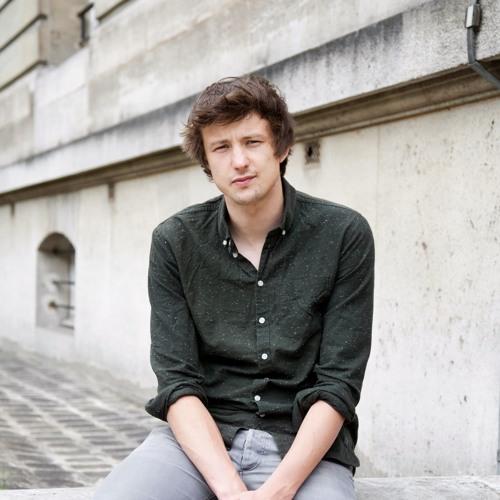 De dichter Bob Vanden Broeck zit voor een grijs gebouw en kijkt in de lens. Bob draagt een donker hemd en een lichtgrijze broek.