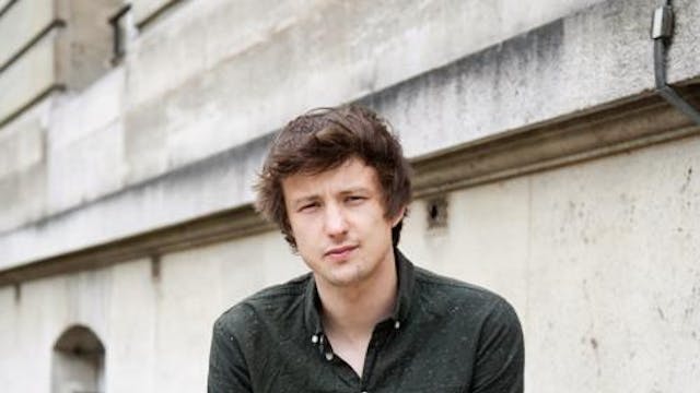 De dichter Bob Vanden Broeck zit voor een grijs gebouw en kijkt in de lens. Bob draagt een donker hemd en een lichtgrijze broek.