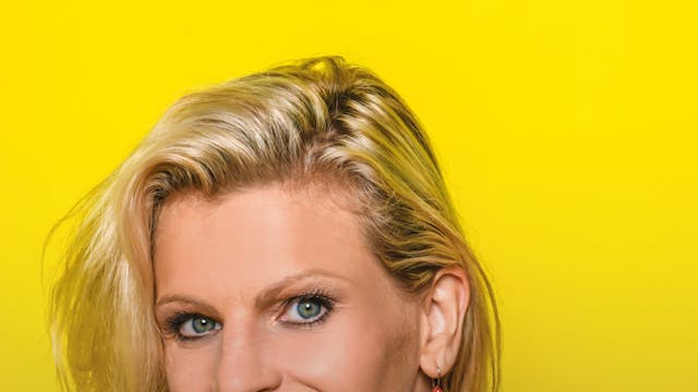 Een vrouw met blond, golvend haar en blauwe ogen, lachend naar de camera, tegen een gele achtergrond. Ze draagt een blauwe top en een opvallende oorbel met het woord "BONUS" erop.