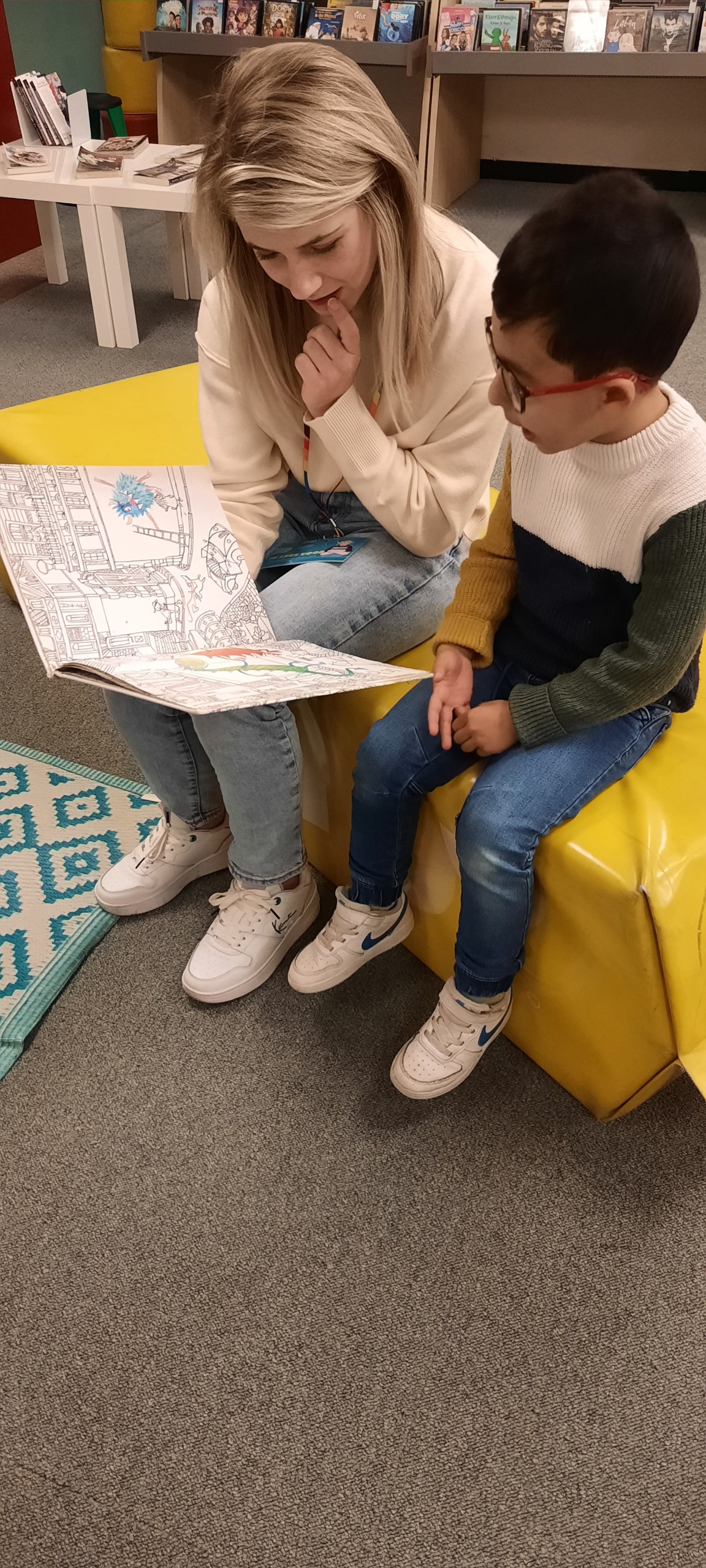 Een vrouw en een kind bekijken samen een kleurboek op een gele bank.